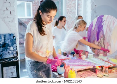 Painting concept. Female paintner artist pouring out yellow paint from one jar to another in art therapy class with two women students finishing picture on background. - Powered by Shutterstock