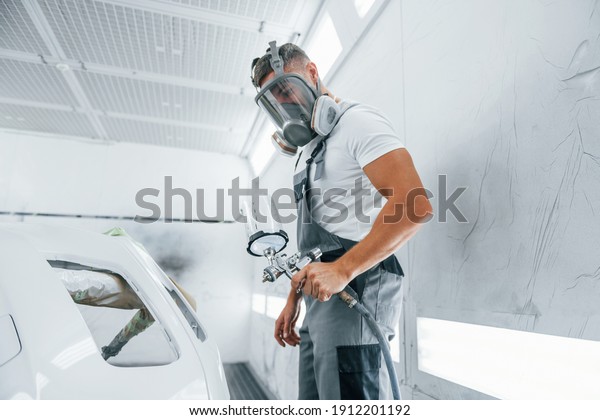 With painting gun. Caucasian automobile repairman\
in uniform works in\
garage.