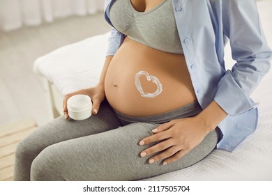 Painting Heart On Pregnant Belly. Pregnant Woman Sitting On Bed With Bare Tummy On Which White Heart Is Made Of Cream. Close Up Of Pregnant Belly Of Woman Holding Container With Stretch Mark Cream.