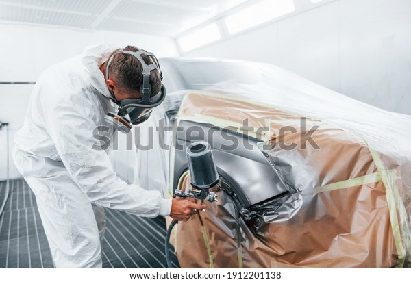 Painting job in progress. Caucasian automobile\
repairman in uniform works in\
garage.