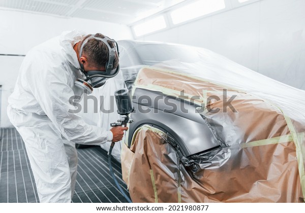 Painting job in progress. Caucasian automobile\
repairman in uniform works in\
garage.