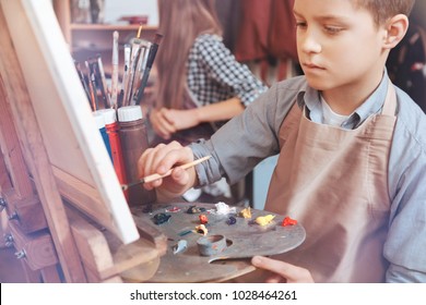Painting is lifefor me. Pensive boy sitting at an easel and looking on a canvas while painting with oil paints during a painting class.