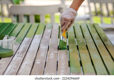 Painting Table With Brush In Protective Gloves. Worker Paints Garden Wood Furniture In Green. Renewing, Renovation Wooden Garden Furniture