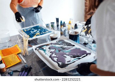 Painting tutorial. Two white women in the gloves paint with liquid acrylic in an art workshop. Acrylic Fluid Pouring. Dirty glass technique. Paintings, paints and brushes on the table. Fluid Art.