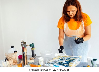 Painting tutorial. White woman in the gloves paint with liquid acrylic in an art workshop. Acrylic Fluid Pouring. Dirty glass technique. Paintings, paints and brushes on the table. Fluid Art.