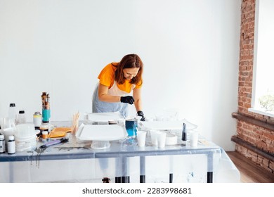 Painting tutorial. White woman in the gloves paint with liquid acrylic in an art workshop. Acrylic Fluid Pouring. Dirty glass technique. Paintings, paints and brushes on the table. Fluid Art.