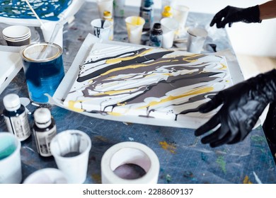 Painting tutorial. Young white girl in the gloves paint with liquid acrylic in an art workshop. Acrylic Fluid Pouring. Dirty glass technique. Paintings, paints and brushes on the table. Fluid Art.