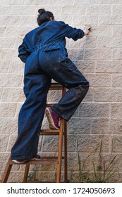 Painting Woman Dressed In Work Overalls Climbing An Old Wooden Ladder Painting With A Paint Brush. Low Angle Shot. Soft Colors.