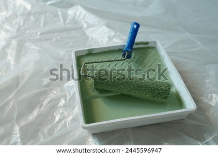 Paintroller with blue plastic handle and green paint in square tray standing on the floor covered with cellophane during renovation work
