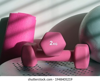 Pair of 2kg pink dumbbells, a rolled yoga or Pilates mat and small grey fitness stability ball. Home gym equipment. Shadows cast by sunlight and a venetian blind. 