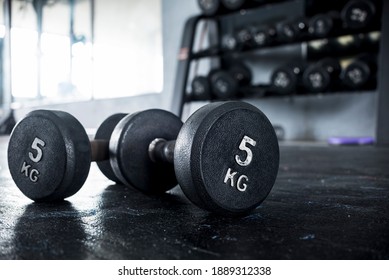 A pair of 5 kilogram dumbbells lying on the floor of a gym, with a dumbbell rack visible in the background.