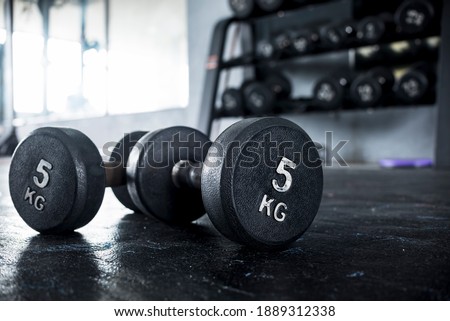 A pair of 5 kilogram dumbbells lying on the floor of a gym, with a dumbbell rack visible in the background.