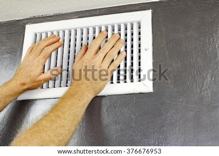 A pair of adult male hands feeling the flow of air coming out of an air vent on a wall near a ceiling. Man with hands in front of an air vent feeling for air flow.