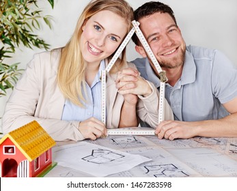 A Pair Of Attractive Blonde Long Haired Women And A Black Haired Handsome Men, Sit Together At The Office Table With Constrtuction Plans And Look Through A House Build With A Ruler