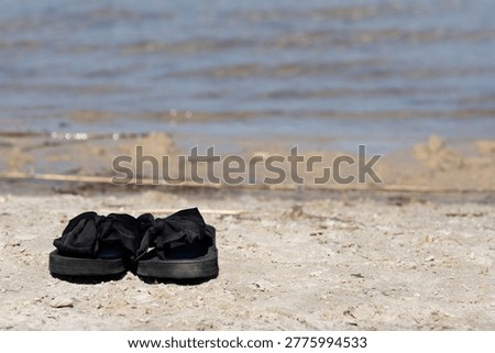 Pair of black women’s flip flops placed near a tranquil lake. Minimalist summer composition with natural light, evoking relaxation, vacation vibes, and a casual lifestyle by the water.