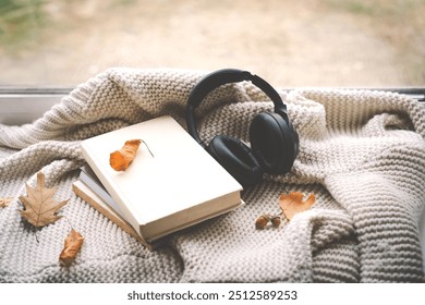 A pair of black headphones lies on an book, surrounded by autumn leaves and a knitted blanket, creating a warm and inviting atmosphere perfect for relaxation. Read, Rest. Cozy autumn or winter concept