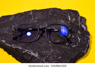 A pair of black rectangular blue light blocking glasses resting on a gray rock against a yellow background.