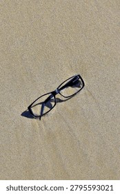 A pair of black rectangular eyeglasses rests lightly on smooth, sunlit sand, casting soft angled shadows. The simple placement and clean texture create a quiet, minimal scene that hints at a moment paused or something left behind.