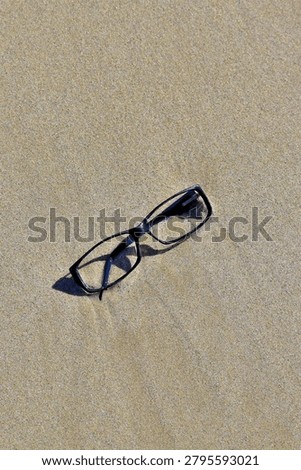 A pair of black rectangular eyeglasses rests lightly on smooth, sunlit sand, casting soft angled shadows. The simple placement and clean texture create a quiet, minimal scene that hints at a moment paused or something left behind.