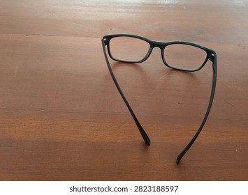 A pair of black-rimmed glasses with rectangular lenses resting on a brown wooden table surface.
