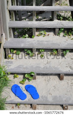 A pair of blue flip flops sit in the sand at the bottom of a set of beach access stairs.