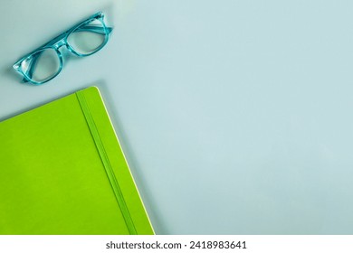 A pair of blue reading glasses and a green notebook on a solid b