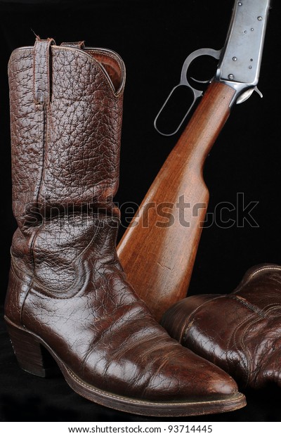 Pair of\
caribou hide cowboy boots and western lever-action rifle against\
black background depicting resting\
wrangler.