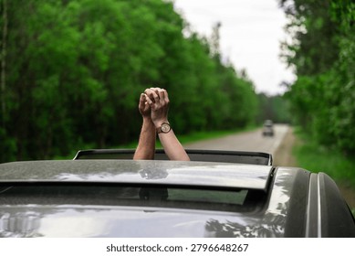 Pair of clasped hands extends through the open sunroof of moving car on tree lined road, with blurred green woodland and an overcast sky in background