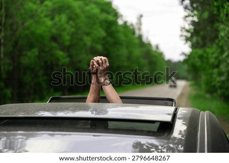 Pair of clasped hands extends through the open sunroof of moving car on tree lined road, with blurred green woodland and an overcast sky in background