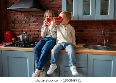 Pair Of Cute Adorable Caucasian Blond Siblings Boy And Girl Enjoy Have Fun Sitting On Kitchen Counter And Drinking Hot Tea Or Chocolate From Big Red Mug. Happy Family Kid And Healthy Lifestyle