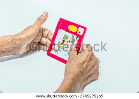 a pair of elderly hands holding a red sliding puzzle toy with animal illustrations on a plain white background, presenting a moment of focus.