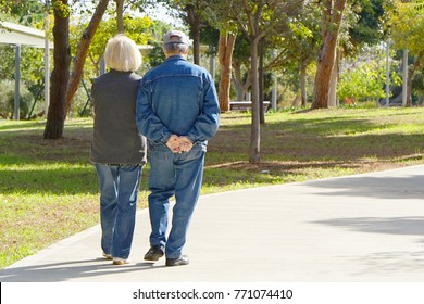 A Pair Of Elderly People Walking In The Park. Back View