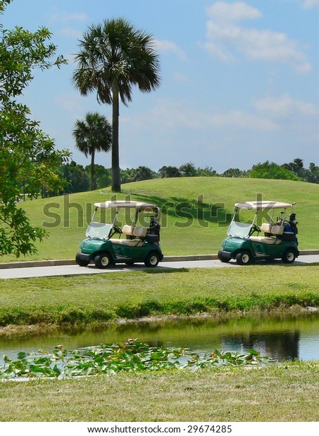 A
pair of electric golf carts parked beside tropical golf course on a
sunny day.  There is a pond in the foreground and a small rolling
green grassy hill. There are few clouds in the blue
sky