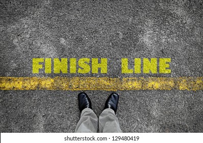 A pair of feet standing at the edge of a yellow line on an asphalt road with a yellow print of the word Finish Line for the concept of accomplishment.