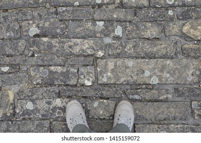 Pair Of Feet In White Canvas Shoes Standing On Grit Stone Sidewalk. Directly Above View. Copy Space. 