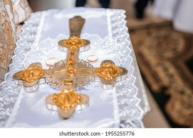 A Pair Of Gold Wedding Rings And An Iron Cross On A White Background In A Church. Wedding Ceremony In The Church