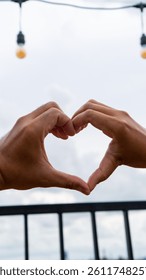 A pair of hands forms a heart shape against a cloudy sky backdrop with string lights, perfect for expressing love and affection.
