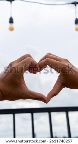 A pair of hands forms a heart shape against a cloudy sky backdrop with string lights, perfect for expressing love and affection.