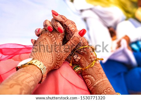 Pair of hands  holding each other  in close up with henna tatoo  with indian designs designs and red nail polish  and wrist watch  in a marriage ceremony background  in selective focus . 