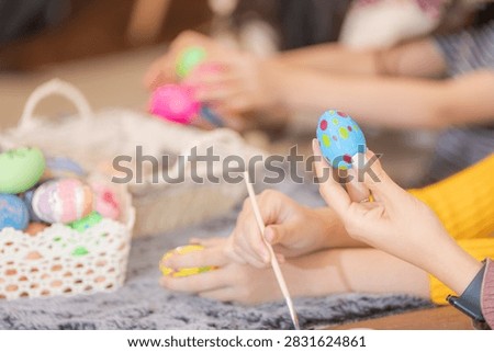 A pair of hands steadying a hand-painted Easter egg above a paint palette on a table covered with paints, brushes and colored eggs at home together as a family on a bright happy day.