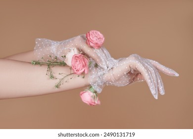 A pair of hands wearing white dotted lace gloves are adorned with pink roses and greenery. The image is shot against a soft brown background. - Powered by Shutterstock