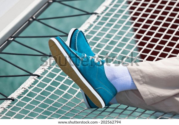 A pair of human legs in\
pants and bright blue topsiders on yacht hammock background.\
Yachting
