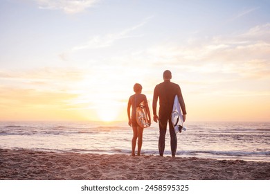 A pair of individuals, son and father in wetsuits stand with surfboards gazing at the sun dipping below the horizon at the beach - Powered by Shutterstock