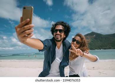 A pair of lovers take a selfie on a tropical beach. The guy and the girl cheerfully smile at the phone camera on the background of the sea. Newlyweds and tropical lagoon with mountains and cloudy sky - Powered by Shutterstock