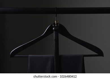 A Pair Of Mens Dark Slacks And Belt Hanging On A Black Hanger And Closet Rod, Set Against A Light To Dark Gray Background.