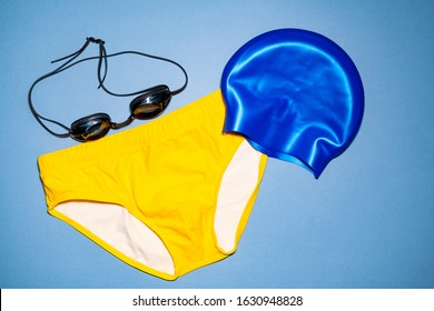 A pair of men's yellow swim briefs, a blue swim cap, and swim goggles on a blue background.