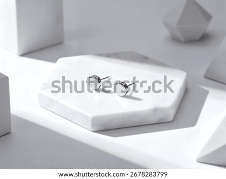 A pair of modern, round silver stud earrings rest on a white marble block with a clean, geometric background.