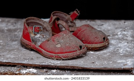A pair of old, red sandals covered in dust are abandoned on a grey surface.