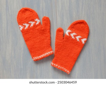 Pair of orange hand knitted mittens decorated with white chevron stripes and Latvian braid on the edge, flatlay on grey background