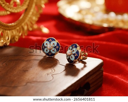 A pair of ornate cloisonné stud earrings, featuring a central pearl surrounded by blue and light blue enamel patterns, resting on a wooden surface with a red fabric background.